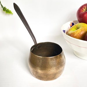 May include: A small, round, brass ladle with a long, black handle. The ladle is sitting on a white surface.