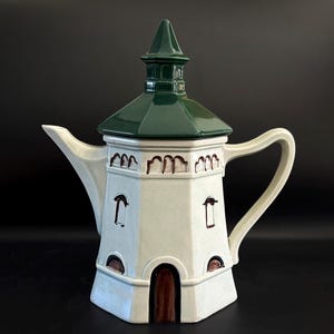 May include: A white ceramic teapot with a green roof and a tower-like design. The teapot has a spout, handle, and a brown door and window details. The teapot is a unique and decorative kitchenware item.