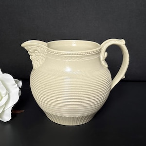 May include: A cream-coloured ceramic pitcher with a ribbed body and a decorative handle. The pitcher has a spout with a sculpted design and a detailed rim. The design is classic and elegant, suitable for serving beverages or as a decorative piece.