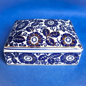 May include: A rectangular, white ceramic box with a blue floral pattern and gold accents. The lid and sides are decorated with stylized flowers and leaves. The box has a gold trim and is set against a blue background.