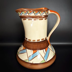 May include: A ceramic jug with a cream-coloured body and a brown base. The jug features geometric patterns in blue, orange, and green. The handle is orange, and the rim has a brown and orange design. This decorative jug is ideal for serving beverages.