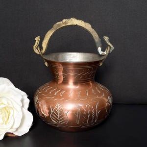 May include: A copper-colored metal vase with a decorative handle. The vase has etched designs and a silver-colored interior. A white flower is in the lower left corner. The handle is gold-colored.