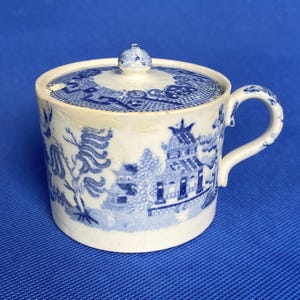 May include: A white ceramic sugar bowl with a blue and white willow pattern. The bowl has a lid and a handle.