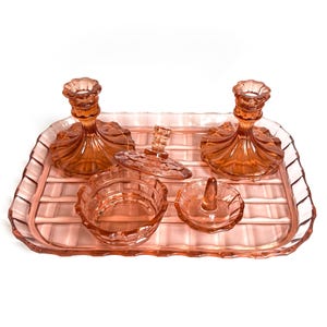 May include: A set of pink depression glass vanity items including a rectangular tray with a grid pattern, two matching candle holders, a small lidded jar, and a ring holder dish, all featuring a translucent, pressed glass construction.