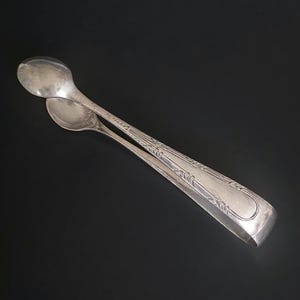 May include: Silver sugar tongs with an ornate design. The tongs are closed and have a spoon-like shape at the ends.