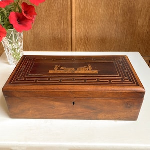 May include: A wooden box with a decorative inlay of a scene with figures and a cart. The box has a dark brown finish and a geometric pattern around the edge.