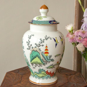 May include: A white porcelain vase with a lid, featuring a hand-painted design of a Chinese landscape with green hills, a red pagoda, and birds in flight. The vase has a blue and gold trim around the lid.