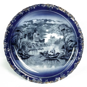 May include: A blue and white porcelain plate with a gold rim. The plate features a transferware design of a river scene with a castle in the background.