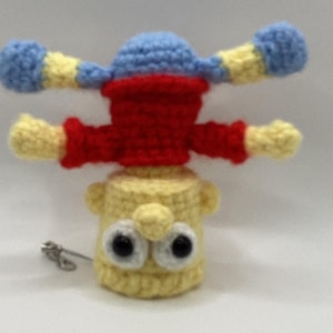 May include: A handmade crochet doll with a yellow body, red shirt, and blue hat. The doll has large black eyes and outstretched arms with blue and yellow accents. A small metal clasp is attached.