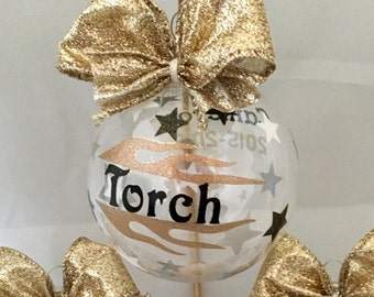 May include: Three clear glass Christmas ornaments with gold glitter bows. Each ornament has a black and gold flame design and the name "Torch" written in black. The ornaments are on a gold stand.
