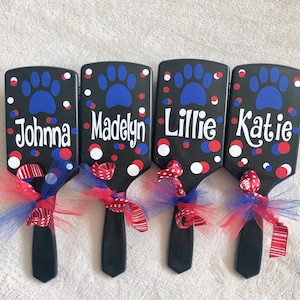 May include: Four black pet brushes with names, paw prints, and red, white, and blue accents. Each brush has a name in white script: Johnna, Madelyn, Lillie, and Katie. The brushes are decorated with red and blue ribbons.