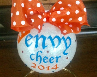 May include: White glitter Christmas ornament with a blue and white "envy cheer 2014" inscription. The ornament is decorated with orange and white polka dot ribbon.