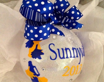 May include: White glitter Christmas ornament with a blue and white polka dot bow. The ornament has a blue and yellow silhouette of a cheerleader and the text "Sunnyvale 2012".