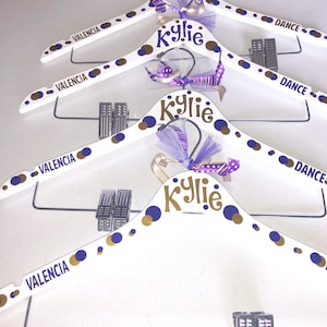 May include: Four white wooden hangers with gold and purple polka dots. Each hanger has the name "Kylie" written in gold letters and the word "Valencia" written in purple letters. The hangers are decorated with purple and gold ribbons.