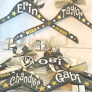May include: Five black wooden hangers with gold stars and white lettering. The hangers are personalized with the names Erin, Taylor, Osi, Chandler, and Gabi. Some of the hangers also have the text "MVAA" and "GO BEARS".