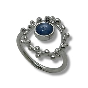 Könnte beinhalten: Ein silberner Ring mit einem blauen Edelstein in der Mitte, umgeben von einem Muster aus kleinen Kugeln. Der Ring hat einen glatten, geschwungenen Band. Das Design schafft eine einzigartige und elegante Ästhetik. Ein Schmuckstück.