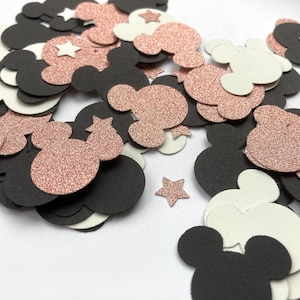 Op de afbeelding: Confetti in de vorm van Mickey Mouse-hoofden en sterren. Het confetti is in zwart, wit en roségoud glitter. Het confetti is verspreid op een witte ondergrond.