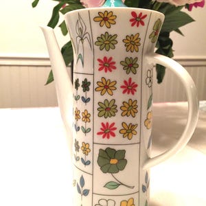 May include: A white ceramic teapot with a tall, slender design. The teapot is decorated with a grid pattern of colorful flowers in yellow, red, and green. It has a teal lid and a curved handle.