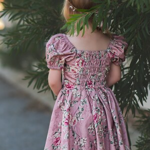 Pu&ograve; includere: Una bambina che indossa un vestito rosa a fiori con maniche a sbuffo e una corona di fiori. Il vestito ha una vita arricciata e una gonna ampia.