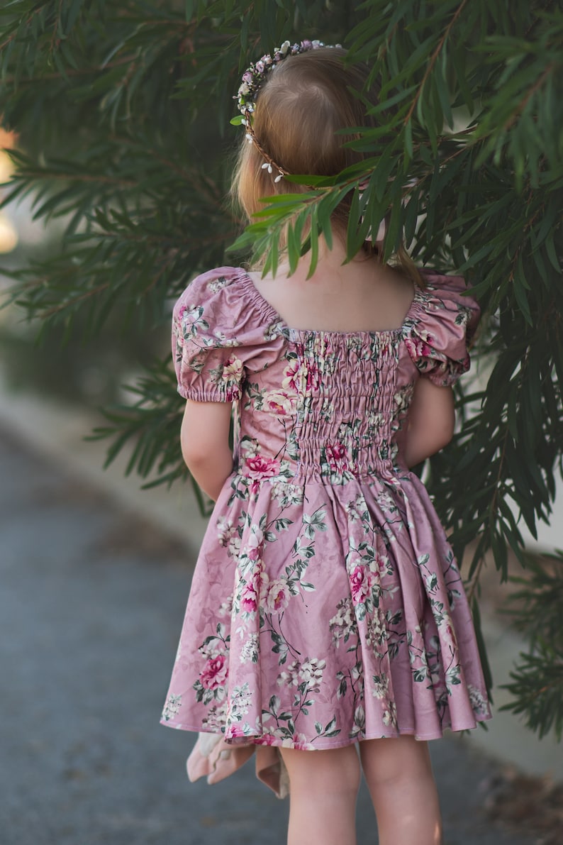 Pu&ograve; includere: Una bambina che indossa un vestito rosa a fiori con maniche a sbuffo e una corona di fiori. Il vestito ha una vita arricciata e una gonna ampia.