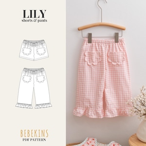 May include: Pink and white gingham shorts and pants pattern, "LILY", hanging on a wooden hanger. The pattern includes sizes 1-12 years. The pattern is from BEBEKINS. The shorts and pants have scalloped pockets and a ruffled hem.