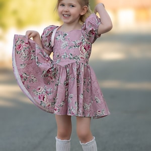 Pu&ograve; includere: Una bambina che indossa un vestito rosa a fiori con un nastro bianco, calze bianche al ginocchio e scarpe marroni. Sorride e ha una corona di fiori in testa.