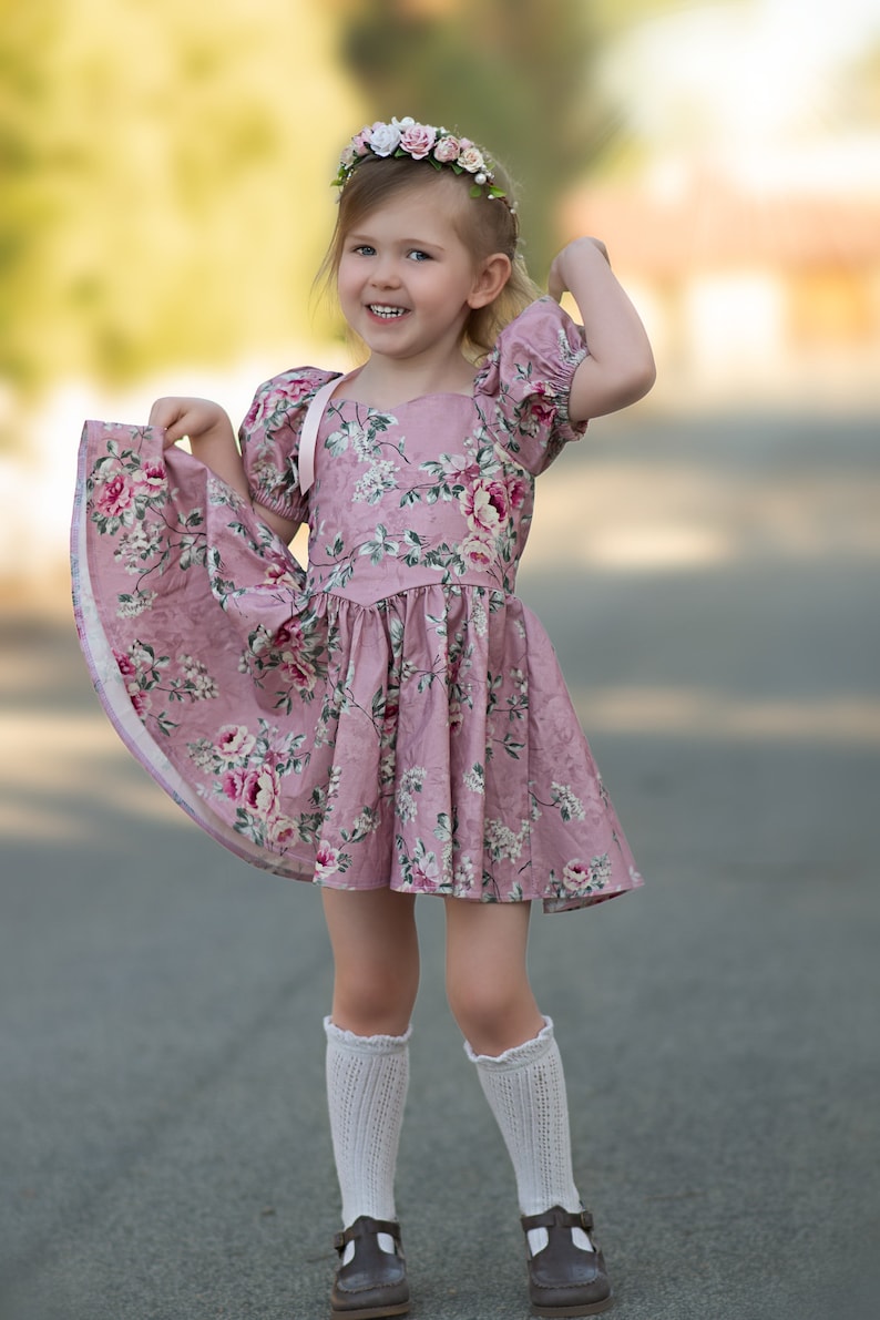 Pu&ograve; includere: Una bambina che indossa un vestito rosa a fiori con un nastro bianco, calze bianche al ginocchio e scarpe marroni. Sorride e ha una corona di fiori in testa.
