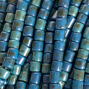 May include: Close-up of a collection of turquoise and teal ceramic beads. The beads are rectangular with rounded edges and a glossy finish. The beads are strung together, creating a textured and colourful pattern.