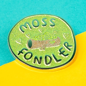 Peut inclure: Une épingle en émail verte et dorée avec le texte "Moss Fondler" et une image de dessin animé d'une bûche mousseuse avec des cœurs.