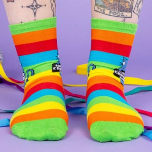 a model with tattooed legs wearing Katie Abey What a Crock of Shit socks with blue cat holding a sign. They are rainbow striped and lovely and vibrant. The model is stood on a yellow floor with balloons, confetti and ribbons