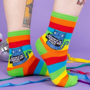 a model with tattooed legs wearing Katie Abey What a Crock of Shit socks with blue cat holding a sign. They are rainbow striped and lovely and vibrant. The model is stood on a yellow floor with balloons, confetti and ribbons