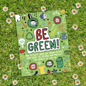 The Be Green Activity Book illustrated by Katie Abey. The book is laid on the grass amongst daisies. The front cover of the book is green with various illustrations of people, animals, symbols, clothes and environmental themes.