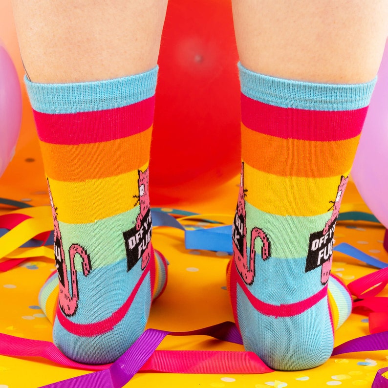 a model with tattooed legs wearing Katie Abey off you fuck socks with pink cat holding a sign. They are rainbow striped and lovely and vibrant. The model is stood on a yellow floor with balloons, confetti and ribbons