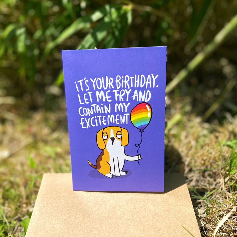 a floppy eared beagle dog on a blue A6 card holding a rainbow balloon looking very unenthusiastic with text saying its your birthday let me try to contain my excitement