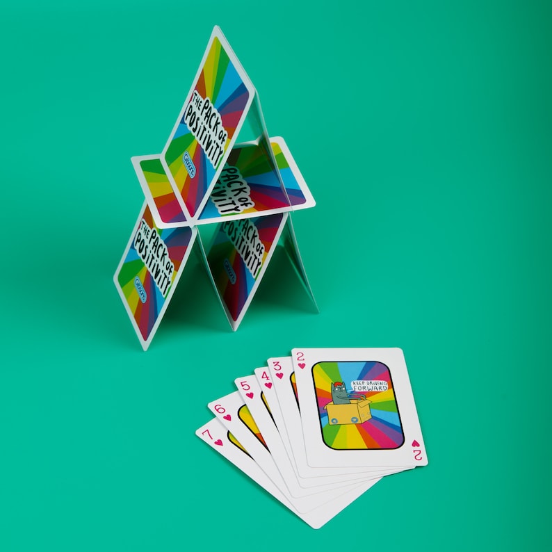 The pack of positivity playing cards in a house formation with a small pile of cards next to it. You can see the rainbow prints and fun illustrated Katie Abey characters on them. They are on a green background.