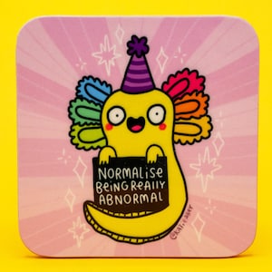 May include: Square coaster featuring a yellow axolotl cartoon character wearing a party hat and holding a sign that reads "Normalise Being Really Abnormal." The background is pink with white star and diamond shapes.