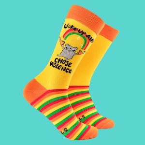 Op de afbeelding: Een paar gele sokken met oranje manchetten en tenen. De sokken hebben een regenboog en een cartoon dier met de tekst "Woke up and Chose Violence". Het onderste deel van de sokken heeft horizontale strepen in rood, oranje, groen en geel.
