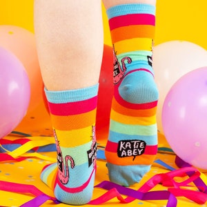 a model with tattooed legs wearing Katie Abey off you fuck socks with pink cat holding a sign. They are rainbow striped and lovely and vibrant. The model is stood on a yellow floor with balloons, confetti and ribbons