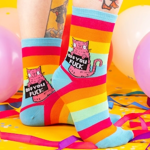 a model with tattooed legs wearing Katie Abey off you fuck socks with pink cat holding a sign. They are rainbow striped and lovely and vibrant. The model is stood on a yellow floor with balloons, confetti and ribbons