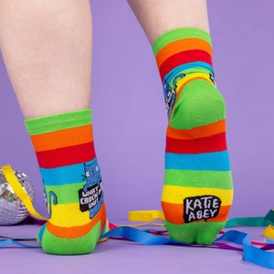 a model with tattooed legs wearing Katie Abey What a Crock of Shit socks with blue cat holding a sign. They are rainbow striped and lovely and vibrant. The model is stood on a yellow floor with balloons, confetti and ribbons