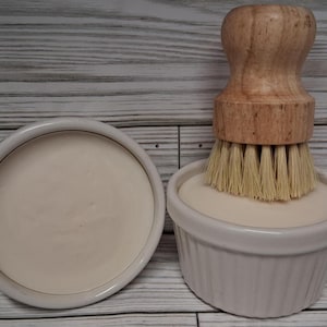 May include: A wooden-handled dish brush with natural bristles rests on a white soap dish. Beside it is a matching dish with a bar of soap. The items are on a light wood-grain surface.