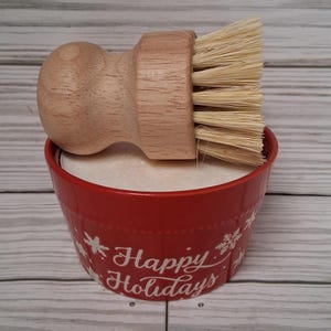 May include: A wooden handled dish brush rests on a red bowl filled with white soap. The bowl has white snowflake designs and the words "Happy Holidays" in white script. The brush has natural bristles.