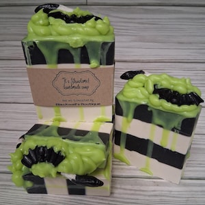 May include: Handmade soap with a layered design of black and white stripes, topped with green soap and black bat accents. The soap has a label that reads "It's Showtime! handmade soap" and "Blackwell's Boutique".