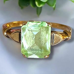 May include: A gold ring with a large, light green, emerald-cut gemstone. The ring has a simple, elegant design.