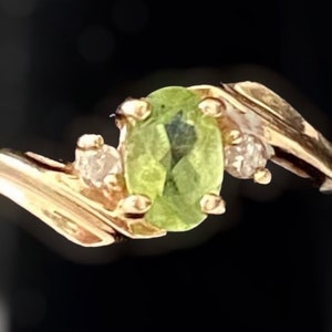 Può includere: Un anello d'oro con una pietra ovale verde e due pietre più piccole trasparenti. L'anello ha una fascia attorcigliata.