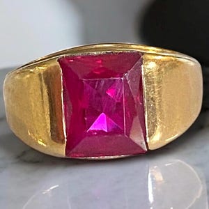 May include: A gold ring featuring a large, square-cut, vibrant red gemstone. The ring has a thick, polished gold band with a classic, bold design. The gemstone is the focal point, catching the light and showcasing its facets.