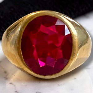 May include: A gold ring featuring a large, faceted, oval-shaped ruby gemstone. The ruby is a vibrant red color, and the gold band has a smooth, polished finish. The ring is a statement piece.