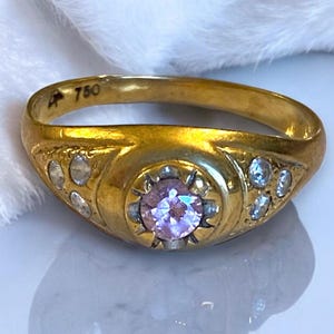 Pu&ograve; includere: Un anello d'oro con una pietra preziosa rosa circondata da pietre preziose pi&ugrave; piccole e trasparenti. L'anello &egrave; contrassegnato con "750" sulla fascia interna.