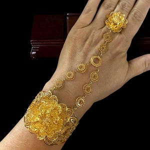 May include: A gold-colored hand chain bracelet with a floral design. The bracelet connects to a ring with a delicate chain. The bracelet features intricate filigree work and is worn on the wrist and hand.