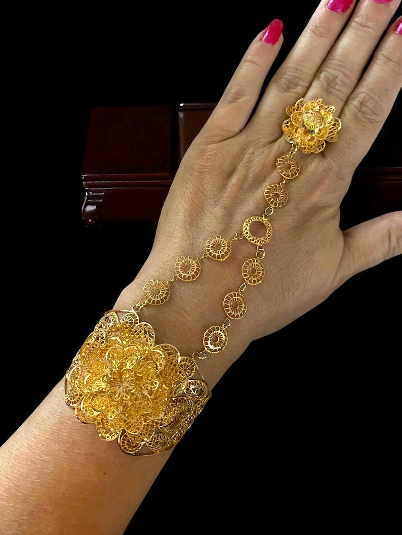 May include: A gold-colored hand chain bracelet with a floral design. The bracelet connects to a ring with a delicate chain. The bracelet features intricate filigree work and is worn on the wrist and hand.
